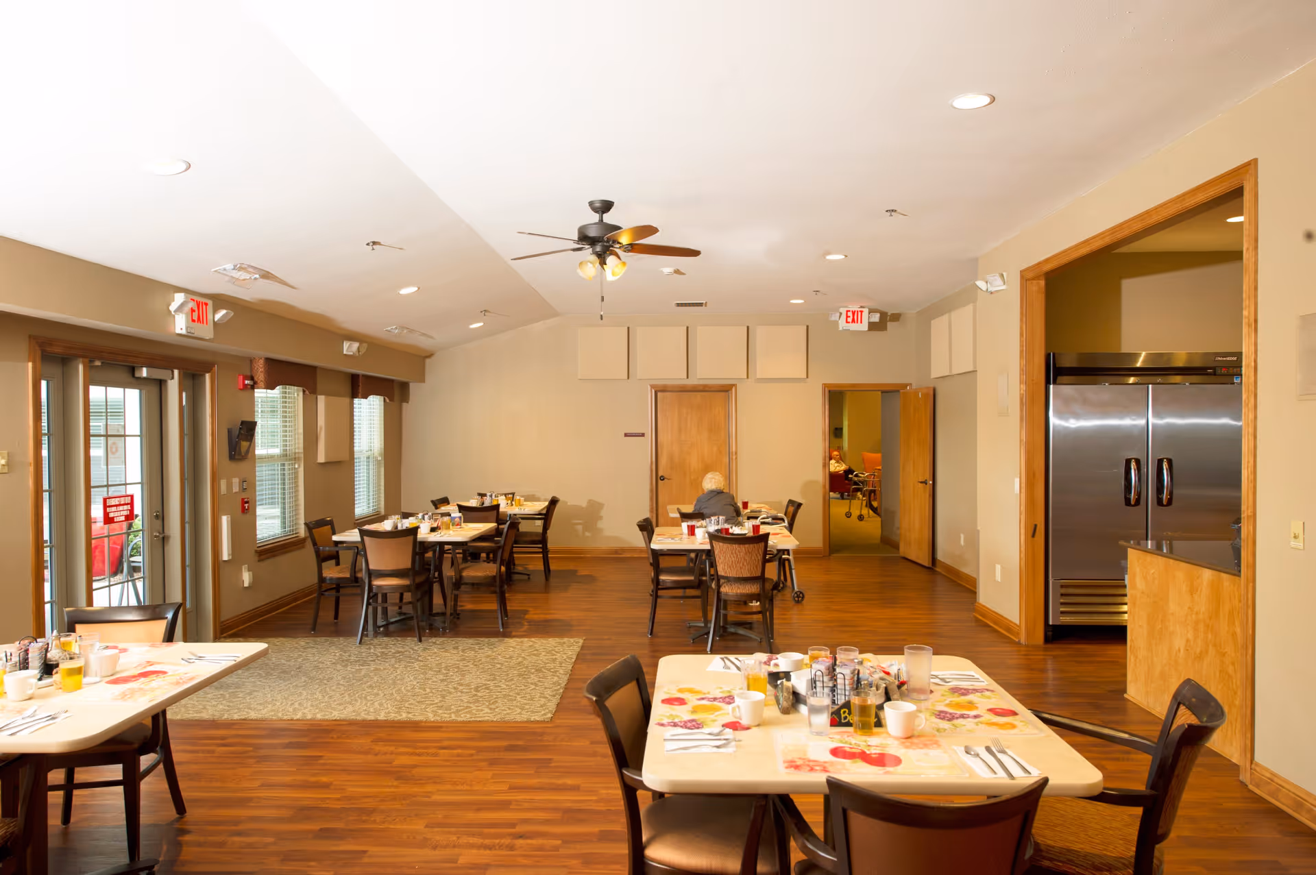 A dining room in a senior living facility with several tables and chairs arranged neatly. Tables are set with placemats, cups, glasses, and utensils. There is a ceiling fan with lights, wooden flooring, and beige walls. A large stainless steel refrigerator is visible through an open doorway on the right. Two exit signs are mounted above doors at the far end of the room. A person with white hair is seated at one of the tables near the back.