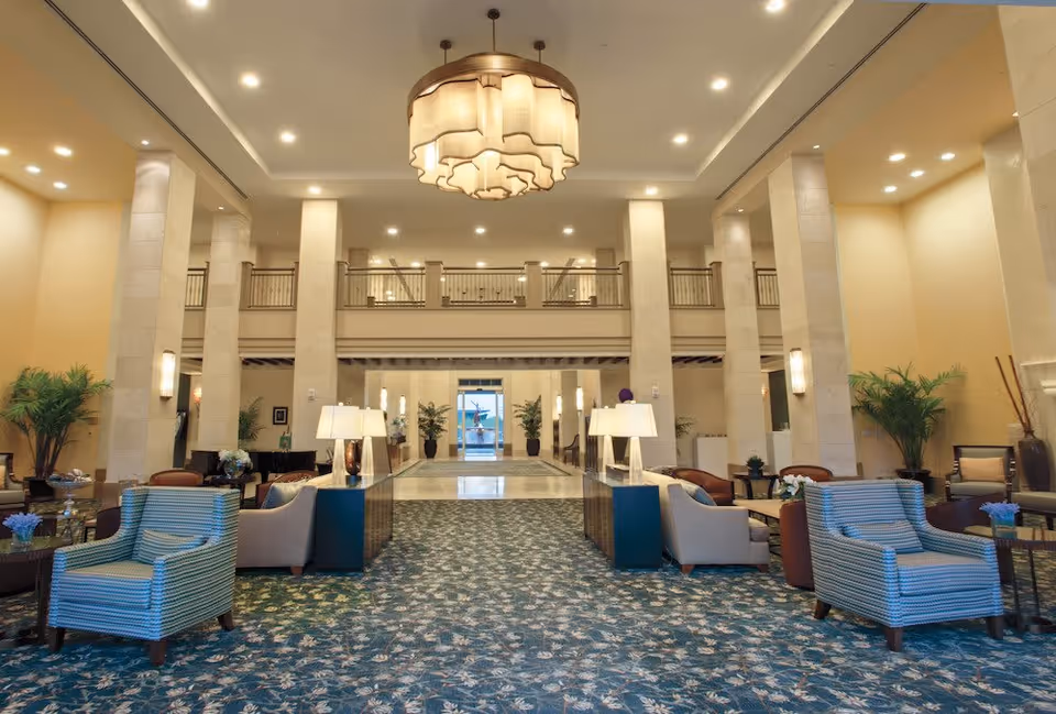 Spacious and well-lit lobby area of a senior living facility with patterned blue carpet, multiple seating arrangements including armchairs and sofas, tall pillars, decorative plants, and a large chandelier hanging from the ceiling.