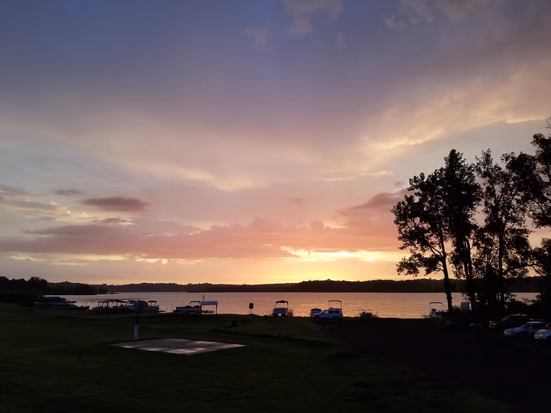 Sunset view over a lake with boats docked along the shore, silhouetted trees on the right, and a grassy area in the foreground.