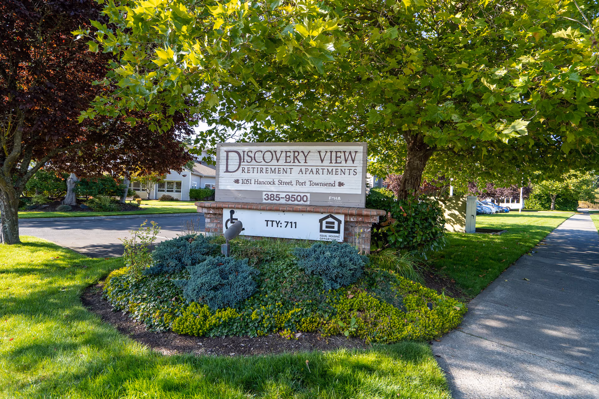 A landscaped entrance sign for Discovery View Retirement Apartments at 1051 Hancock Street, Port Townsend, surrounded by green grass, bushes, and trees with a sidewalk on the right side.