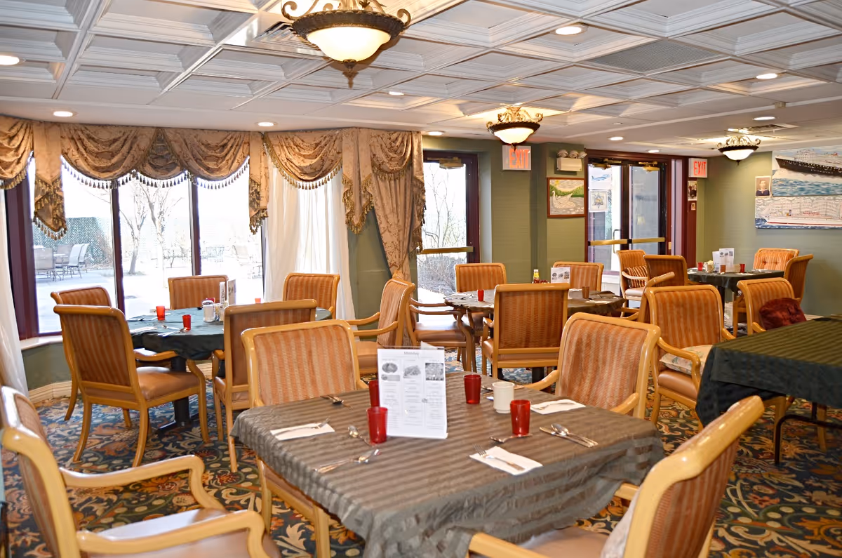 Dining room with multiple tables and cushioned chairs set for meals beneath decorative ceiling lights and large draped windows.