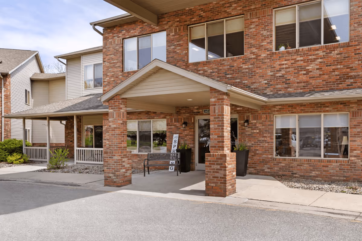 Exterior view of a senior living facility entrance with a covered porch supported by brick columns. There is a bench and two large planters with greenery near the entrance door. The building is made of red brick with multiple windows, and a sign leaning against the wall reads 'SPRING'.