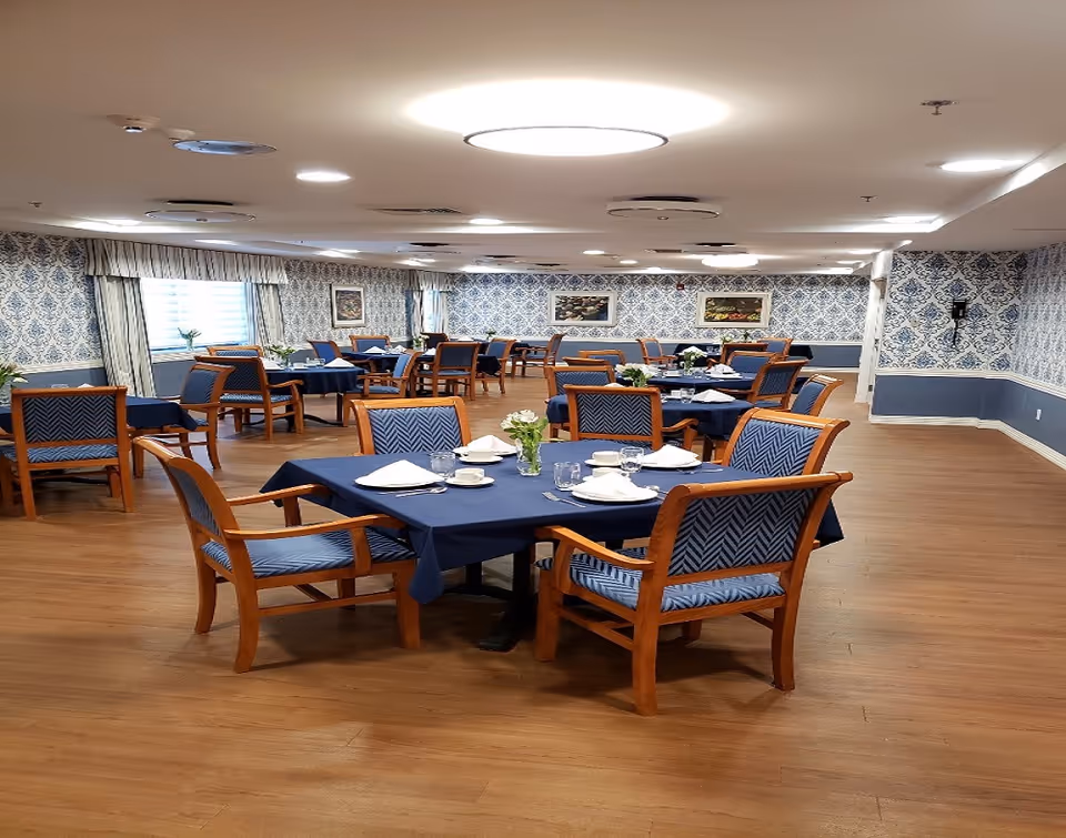 A spacious dining room with multiple wooden tables covered with dark blue tablecloths. Each table is surrounded by wooden chairs with blue patterned cushions. The tables are set with white napkins, cups, glasses, and small flower arrangements. The room has wooden flooring, patterned blue and white wallpaper, and framed artwork on the walls. Ceiling lights provide bright illumination.