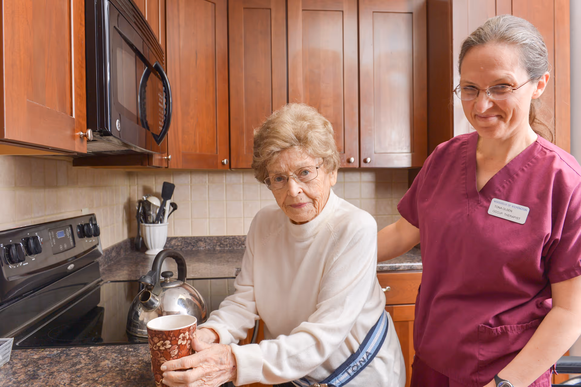 An elderly woman holding a cup in a kitchen with wooden cabinets and a kettle on the stove, accompanied by a smiling caregiver wearing a maroon uniform and glasses.