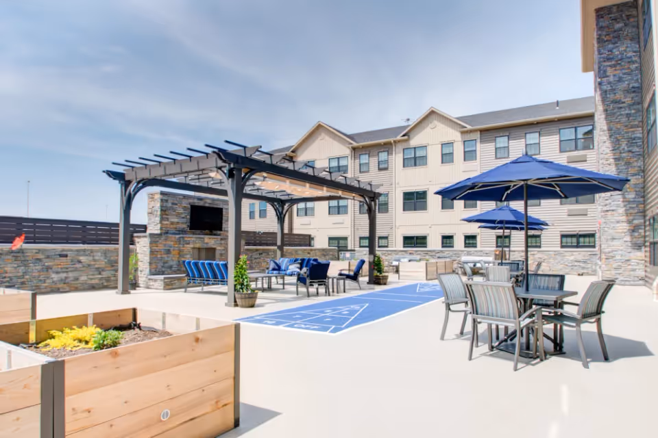 Outdoor courtyard with a pergola, lounge seating, a shuffleboard court, tables with umbrellas, and raised planters beside a multi-story residential building.
