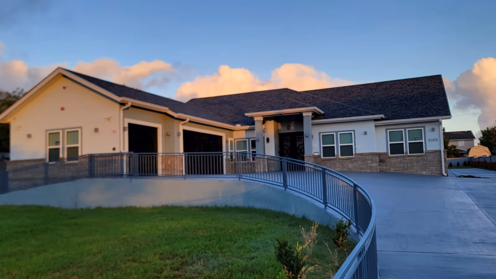 Exterior view of a single-story assisted living facility building with a sloped roof, multiple windows, and a ramp with metal railings leading to the entrance. The building is surrounded by a green lawn and a concrete driveway under a partly cloudy sky during sunset.