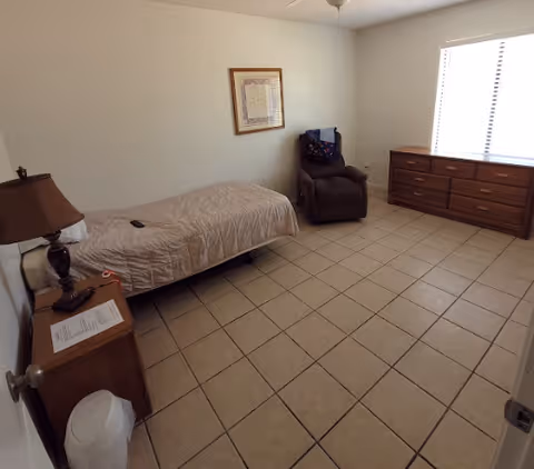 A simple bedroom with a single bed covered with a beige quilt, a brown armchair with a colorful blanket draped over the back, a wooden dresser with multiple drawers under a window with blinds, a wooden nightstand with a lamp and some papers, and a tiled floor.