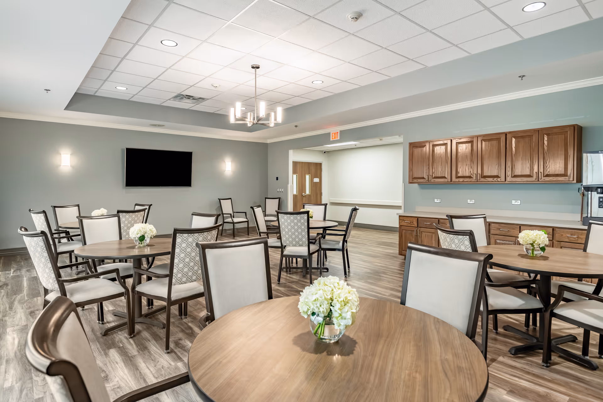 A spacious dining room with multiple round wooden tables, each surrounded by cushioned chairs. Small flower arrangements are placed on each table. The room has wooden flooring, light gray walls, a mounted flat-screen TV, and wooden cabinets along one wall. Ceiling lights and a modern chandelier provide illumination.