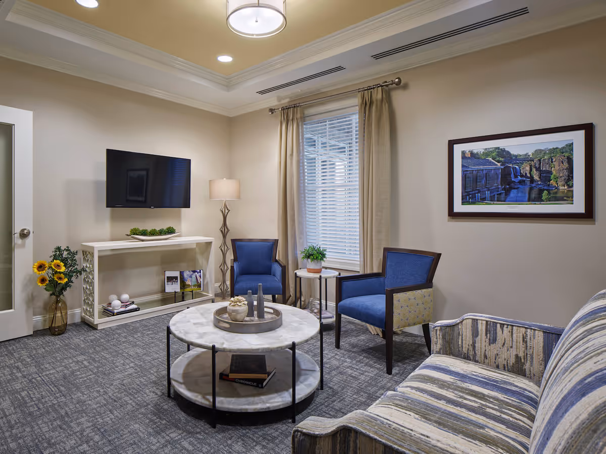 A cozy living room with a striped sofa, two blue armchairs, a round marble coffee table with decorative items, a small side table with a plant, a floor lamp, a wall-mounted flat screen TV, a white console table with decorative items, and a framed landscape picture on the wall. The room has beige walls, a window with beige curtains, and a gray carpeted floor.