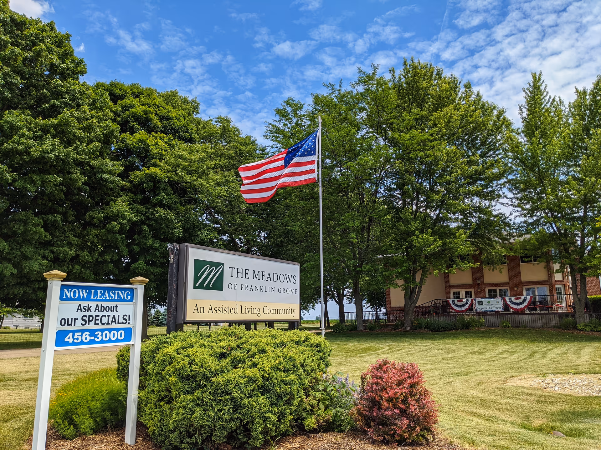 Outdoor view of The Meadows of Franklin Grove Assisted Living community with a large sign displaying the facility's name and description. An American flag is flying on a flagpole behind the sign. There is a smaller sign advertising leasing specials with a phone number. The background shows a brick building partially obscured by trees and a well-maintained lawn.