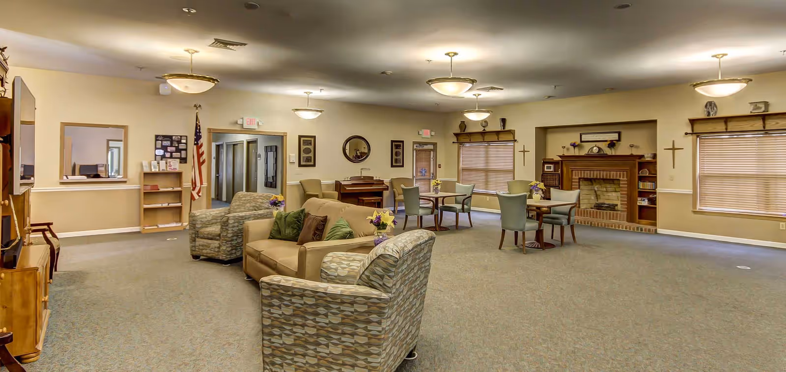 A spacious senior living facility common area with beige walls and carpeted floor. The room features a beige sofa with green and brown pillows, two patterned armchairs, several small round tables with chairs, a wooden fireplace with shelves on either side, and a piano against the far wall. There are multiple ceiling lights, windows with blinds, and an American flag near a doorway.