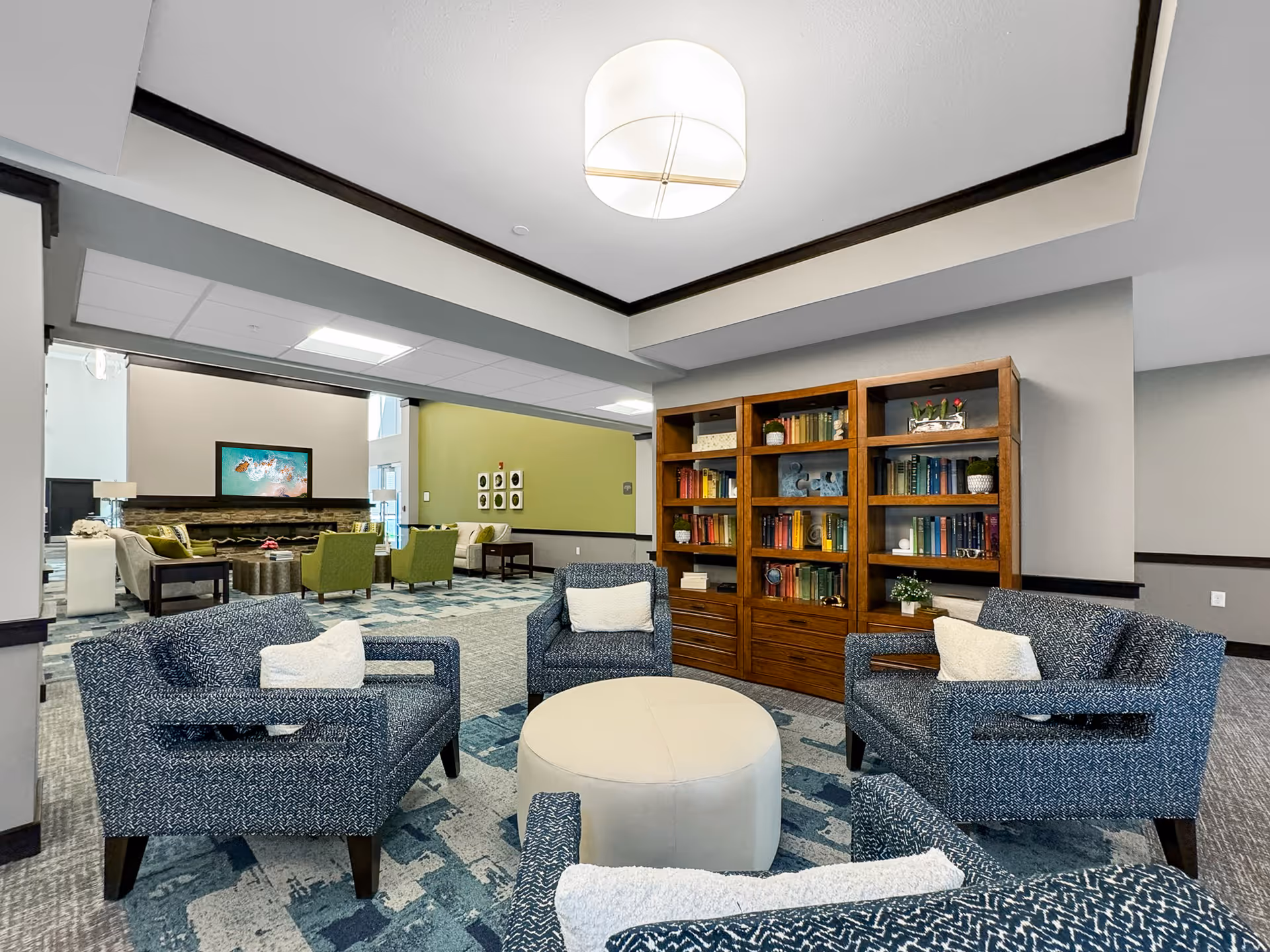 A spacious senior living facility common area with four blue patterned armchairs arranged around a round beige ottoman. Behind the chairs is a wooden bookshelf filled with books and decorative items. In the background, there is another seating area with green chairs and beige sofas, a large wall-mounted TV, and a fireplace. The room features a modern ceiling light fixture and a mix of carpet patterns in blue and gray tones.