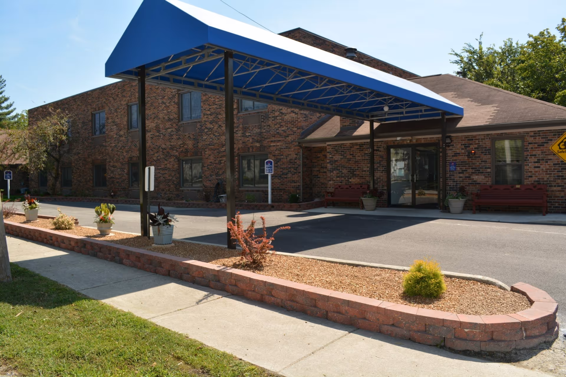 Brick building front with a blue canopy over the driveway, benches by the entrance and planters along a low brick wall.
