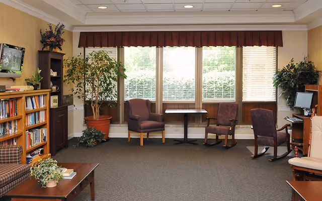 A cozy living room area with a carpeted floor, several upholstered chairs, a small table, a wooden bookshelf filled with books, a wall-mounted TV, and large windows with blinds and a valance. There are also potted plants and a computer desk with a monitor on the right side.