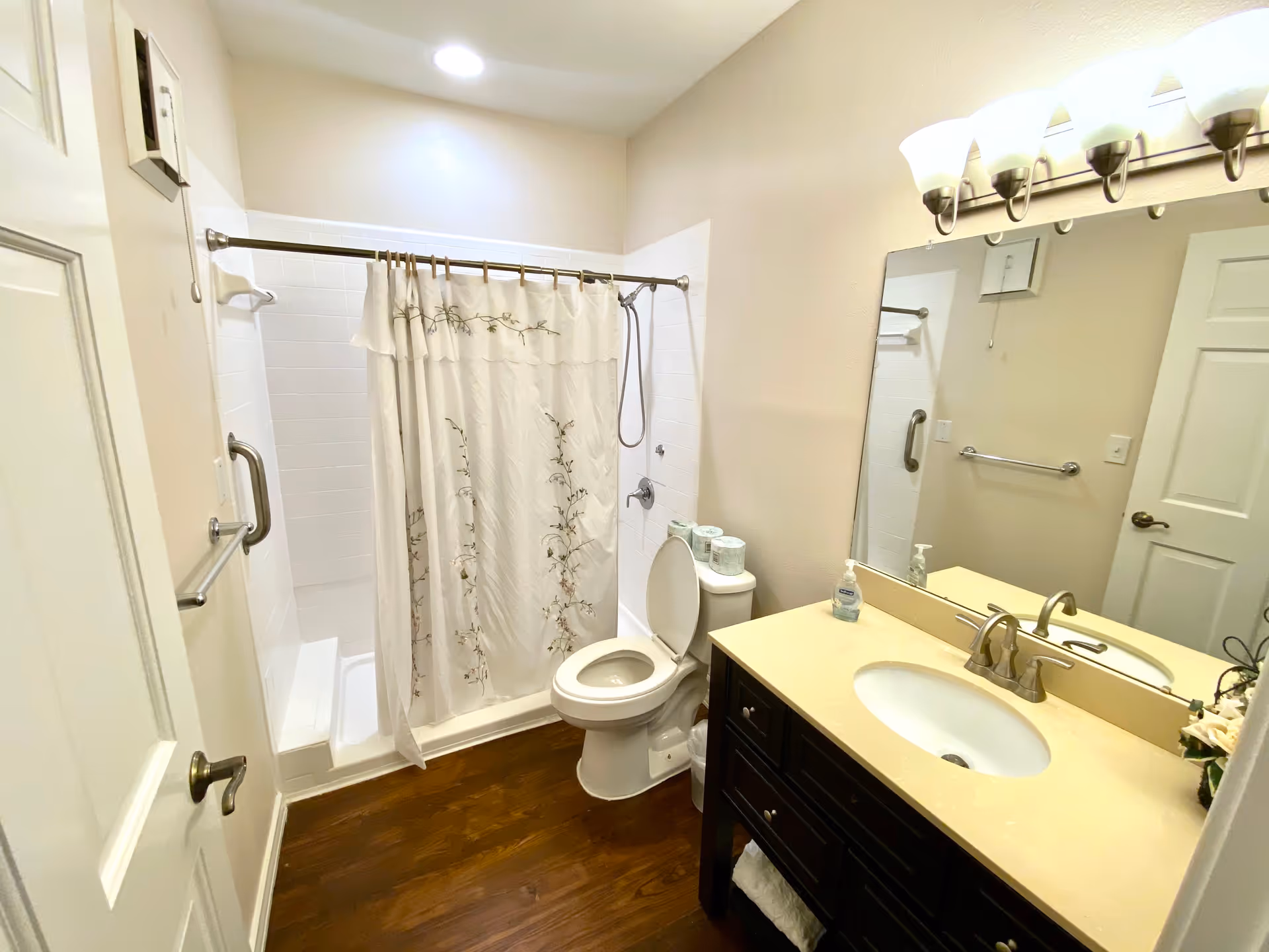 A clean bathroom with a white toilet, a shower with a floral-patterned curtain, a dark wood vanity with a beige countertop and an oval sink, a large mirror above the sink, and a light fixture with multiple bulbs. The floor is wooden, and the walls are painted light beige.
