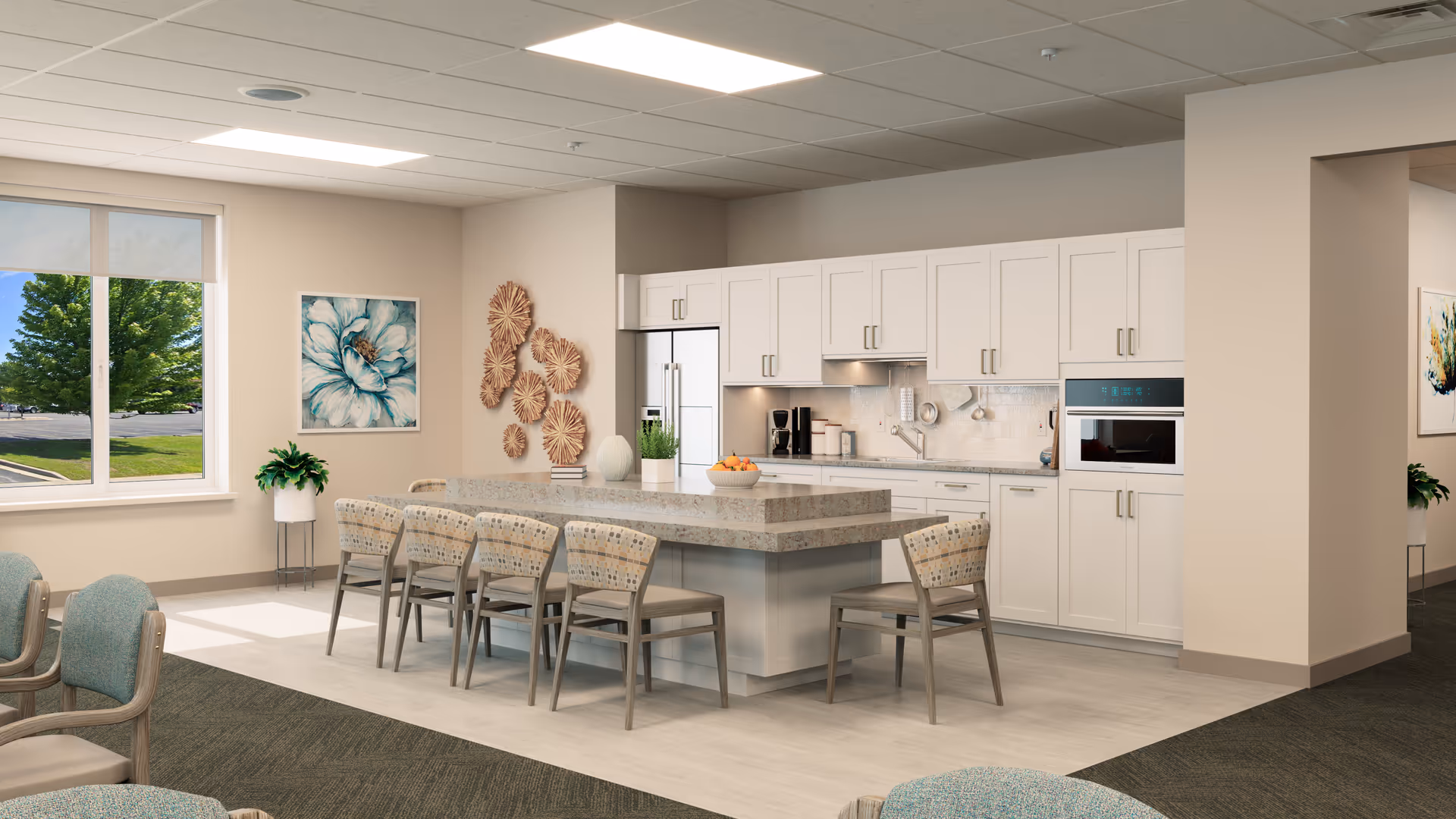 A bright and modern kitchen area in a senior living facility featuring a large island with a granite countertop surrounded by six chairs. The kitchen has white cabinets, built-in appliances including a refrigerator and oven, and a coffee maker on the counter. There is a large window letting in natural light, decorative wall art, and a plant near the window.