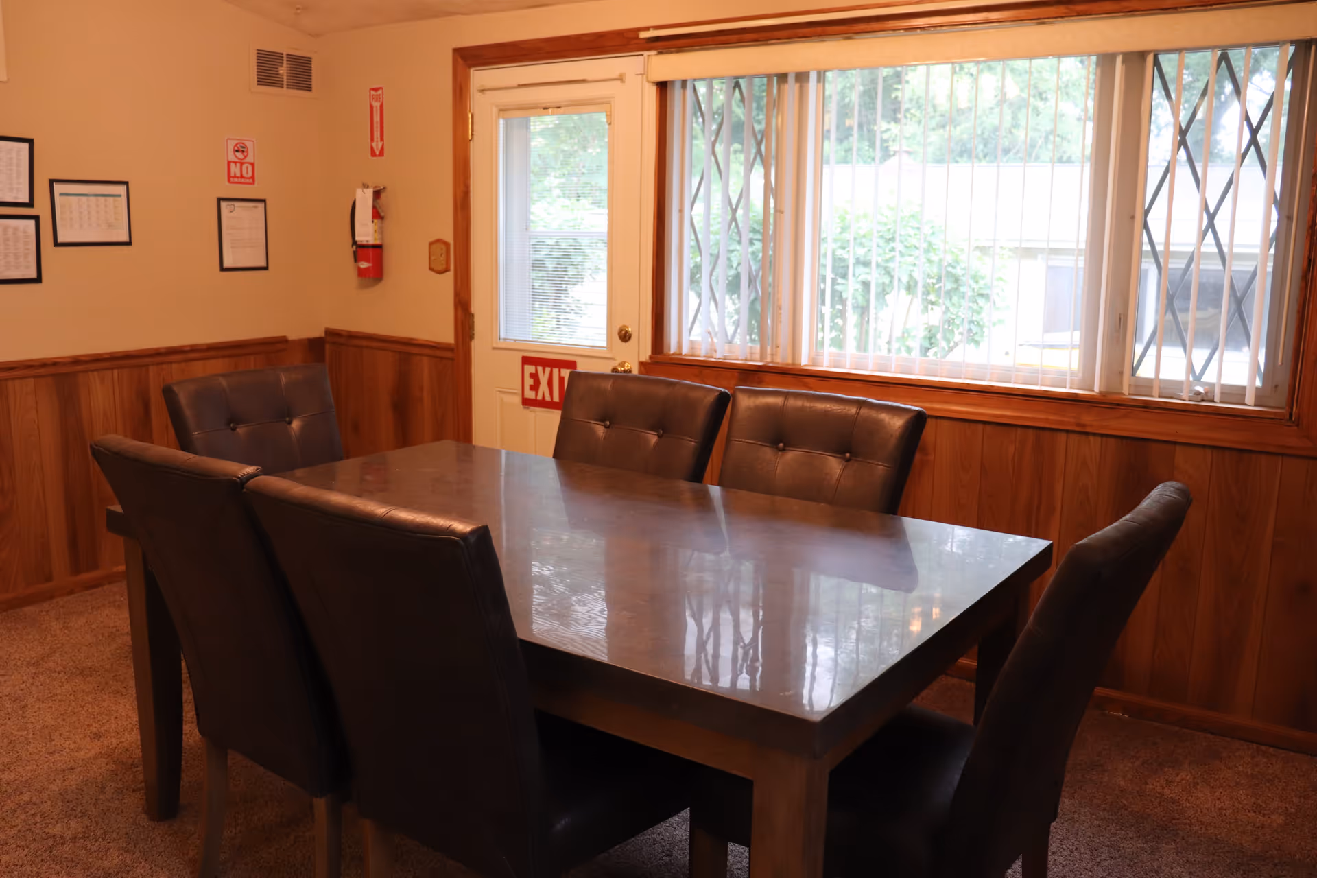 A dining room with a rectangular table and six dark cushioned chairs. The room has wood-paneled walls, a window with vertical blinds, and a door with an exit sign. There are framed documents and a fire extinguisher mounted on the wall.