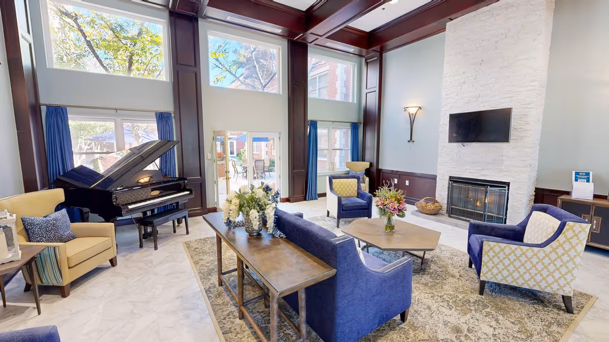 A bright and spacious living room area with large windows and glass doors letting in natural light. The room features a grand piano, several upholstered armchairs and sofas in blue and yellow tones, a wooden coffee table with a flower vase, a fireplace with a mounted flat-screen TV above it, and a decorative rug on the floor. Outside the glass doors, there is a patio area with tables and chairs.