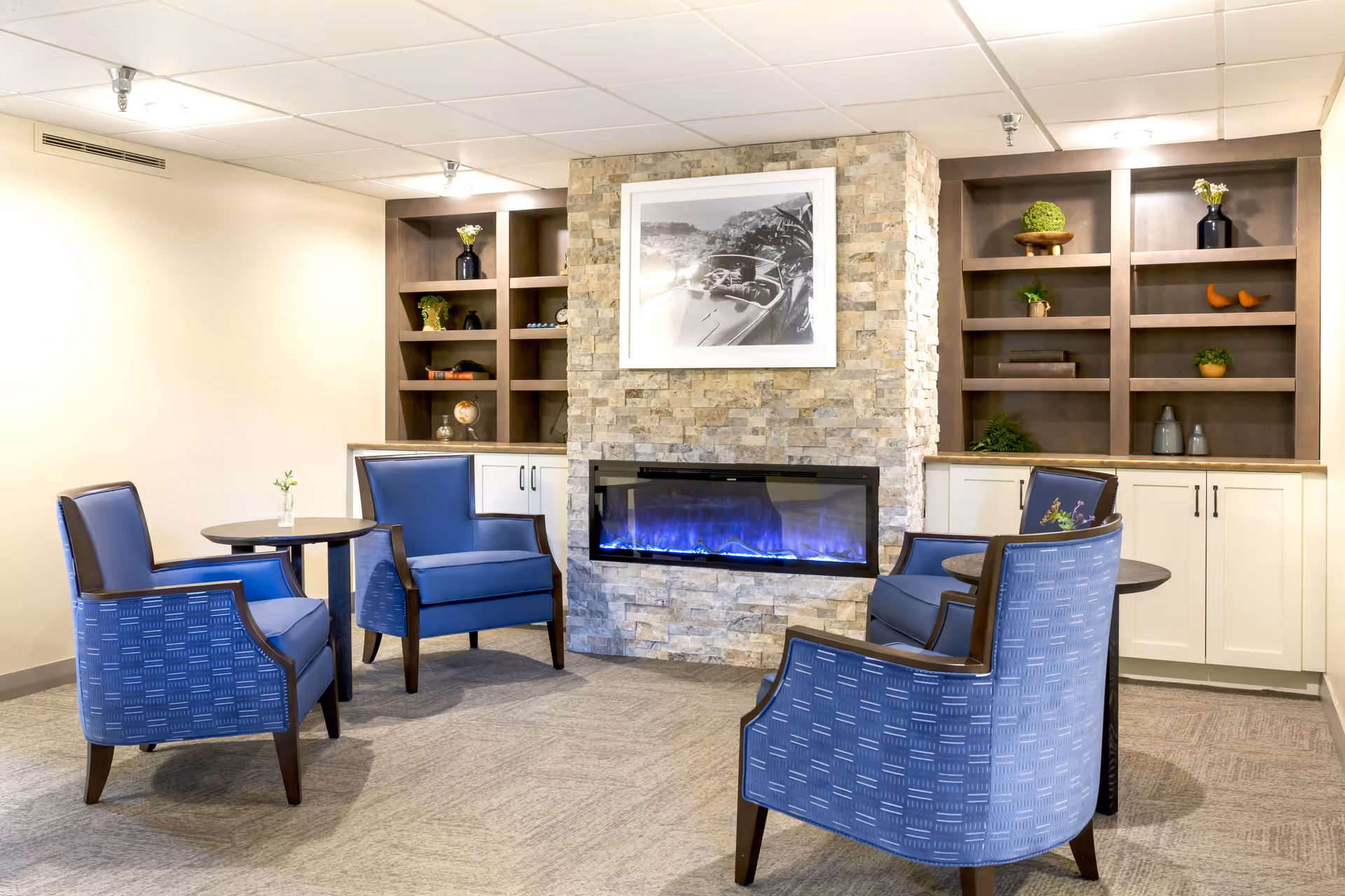 A cozy sitting area with four blue upholstered armchairs arranged around two small round tables. The space features a modern electric fireplace set in a stone wall with built-in wooden shelves on either side, decorated with plants, books, and decorative items. Above the fireplace is a black and white framed photograph.