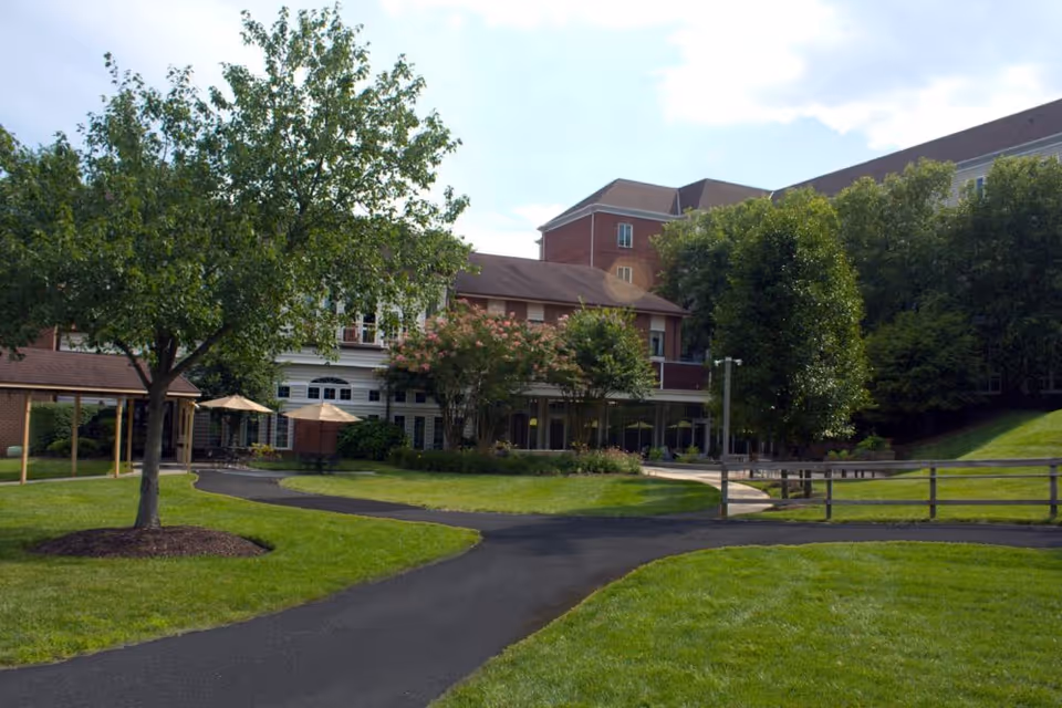 View of a senior living facility's outdoor area with a paved walkway, green grass, trees, and a multi-story building in the background under a partly cloudy sky.