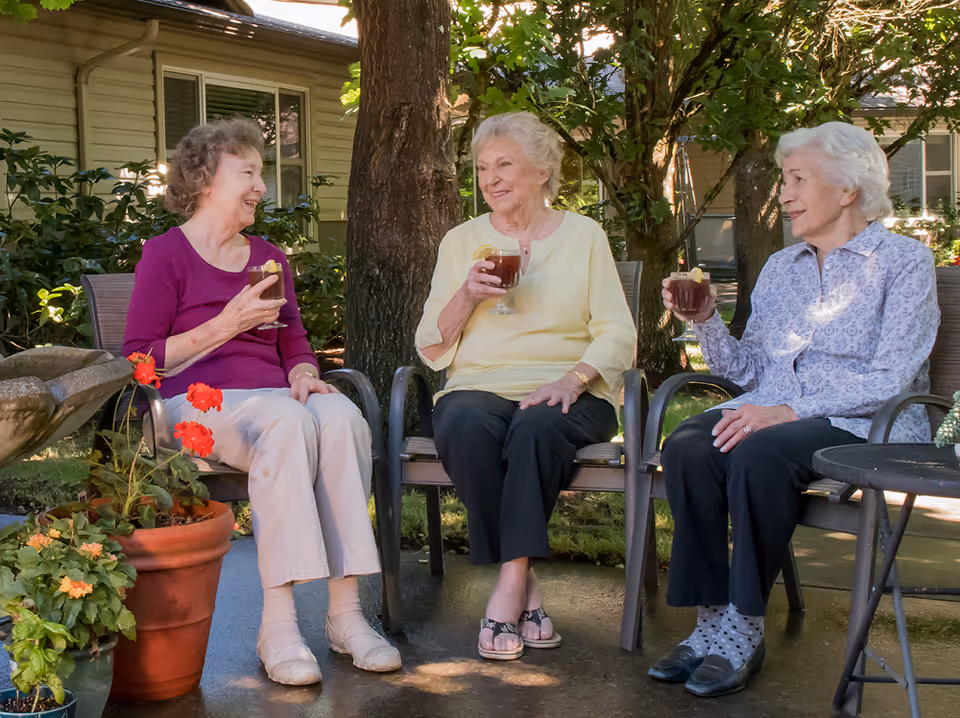Three elderly women sitting outdoors on chairs around a small table, enjoying drinks with lemon slices, surrounded by greenery and potted plants in a garden area of a senior living facility.