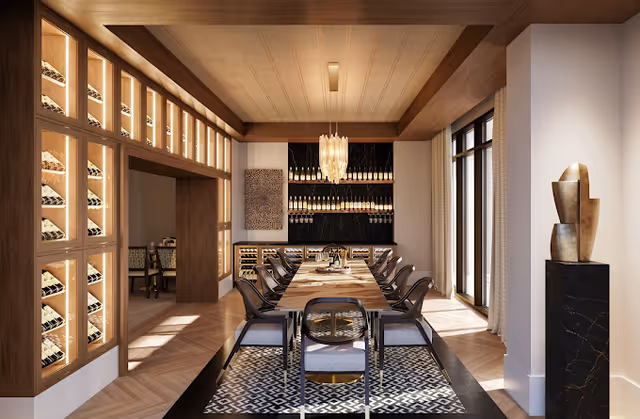 A modern dining room with a long wooden table surrounded by chairs. The room features built-in wine racks filled with bottles on the left wall and a wine display on the far wall. A chandelier hangs above the table, and large windows on the right side allow natural light to fill the space. A decorative sculpture is placed on a pedestal near the windows.