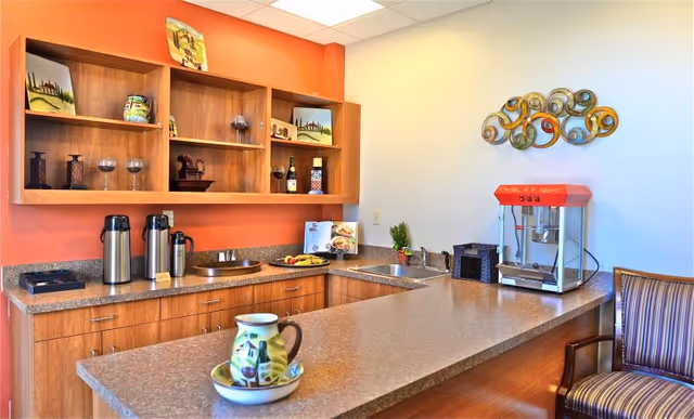 A kitchenette area with wooden cabinets and shelves, a countertop with a decorative pitcher and bowl, coffee dispensers, a popcorn machine, and a striped armchair. The wall behind the counter is painted orange and white, with decorative items on the shelves and a metal wall art piece above the popcorn machine.
