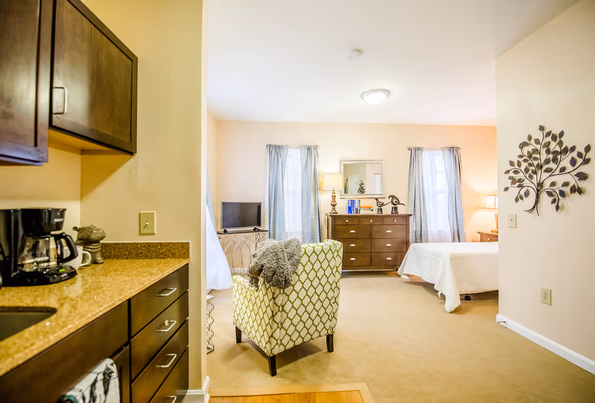 A bright and cozy studio apartment interior with a small kitchen area on the left featuring dark wood cabinets and a coffee maker on the countertop. The main living space includes a patterned armchair with a gray throw draped over it, a wooden dresser with decorative items and books, a small TV on a stand, and a bed with a white quilt. Light blue curtains frame two windows, and a metal wall decoration shaped like a tree is mounted on the right wall.