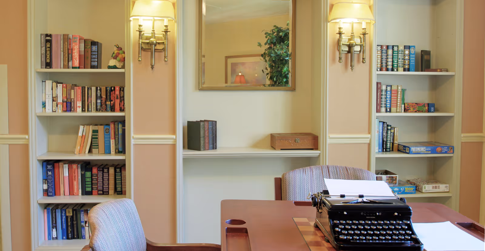 Cozy interior room with built-in bookshelves, a table holding a vintage typewriter, upholstered chairs, wall sconces, and a mirror.