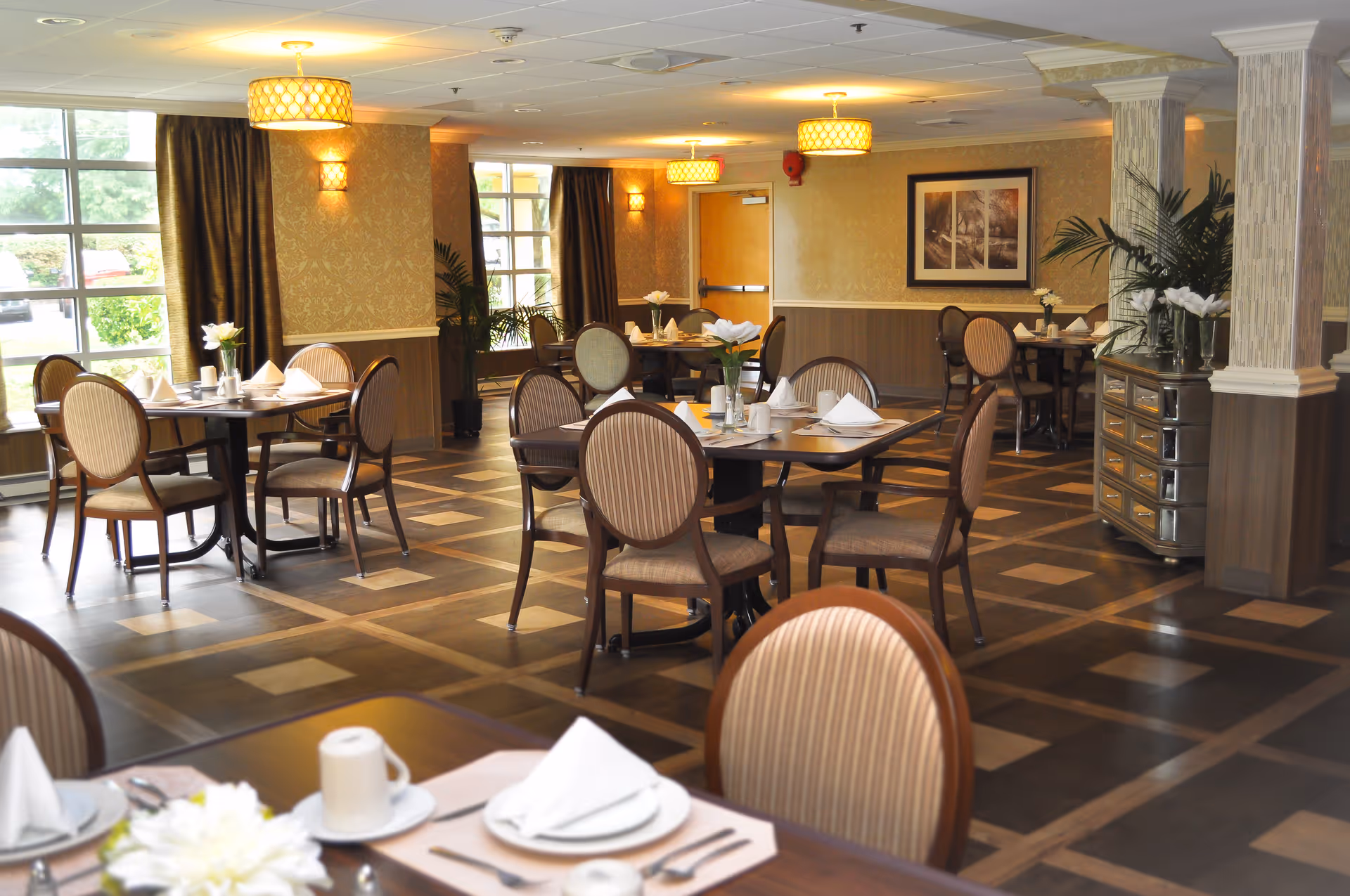 A well-lit dining room with multiple round tables set with white napkins, plates, cups, and silverware. The room features patterned wallpaper, large windows with dark curtains, decorative plants, and framed artwork on the walls. The floor has a geometric wood pattern, and there are several ceiling light fixtures providing warm lighting.