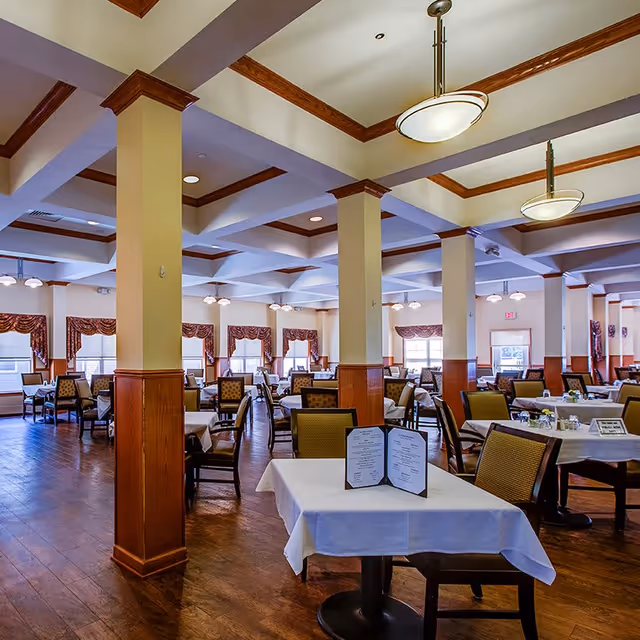 Spacious dining room with multiple tables covered in white tablecloths, surrounded by cushioned chairs. The room features wooden flooring, cream-colored walls with wooden paneling on the lower half, large windows with patterned valances, and ceiling lights hanging from a coffered ceiling.
