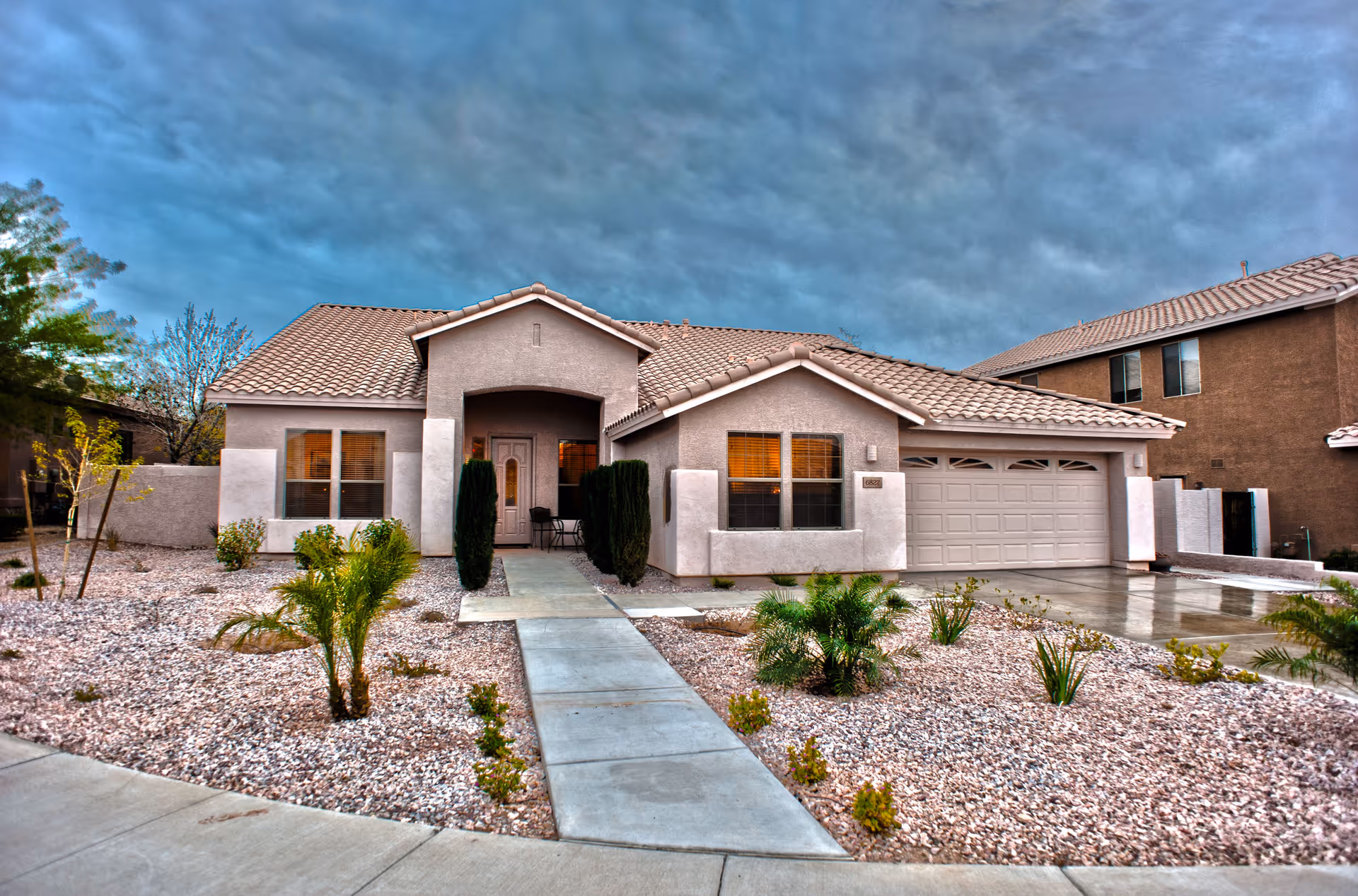 Front exterior view of a single-story house with a tiled roof, beige stucco walls, a concrete walkway leading to the front door, and a two-car garage. The front yard is landscaped with rocks, small plants, and shrubs under a cloudy sky.