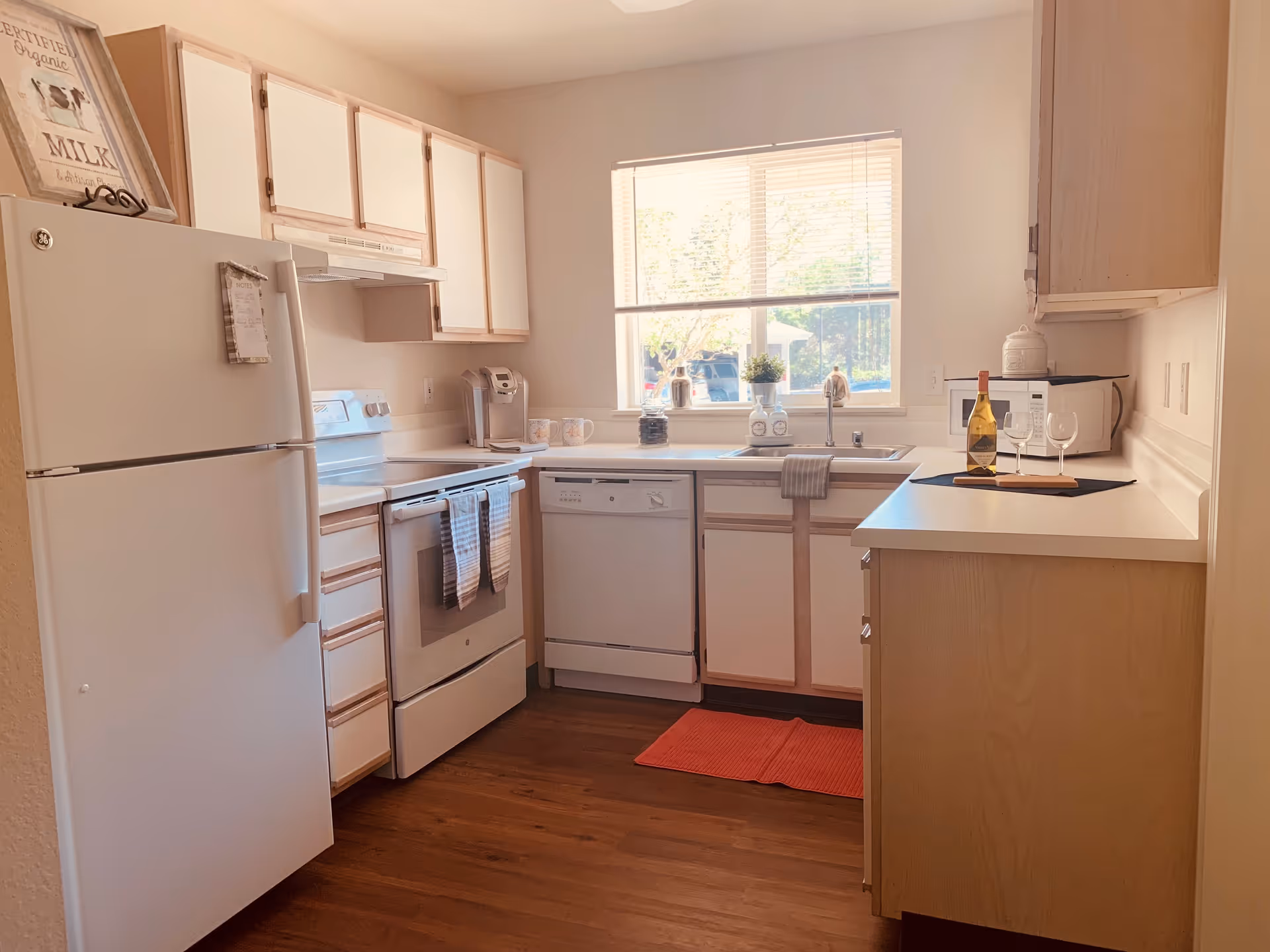 A bright kitchen with white cabinets and appliances including a refrigerator, stove, dishwasher, and microwave. There is a window above the sink letting in natural light. A red rug is on the wooden floor, and a bottle of wine with two glasses is placed on the counter.