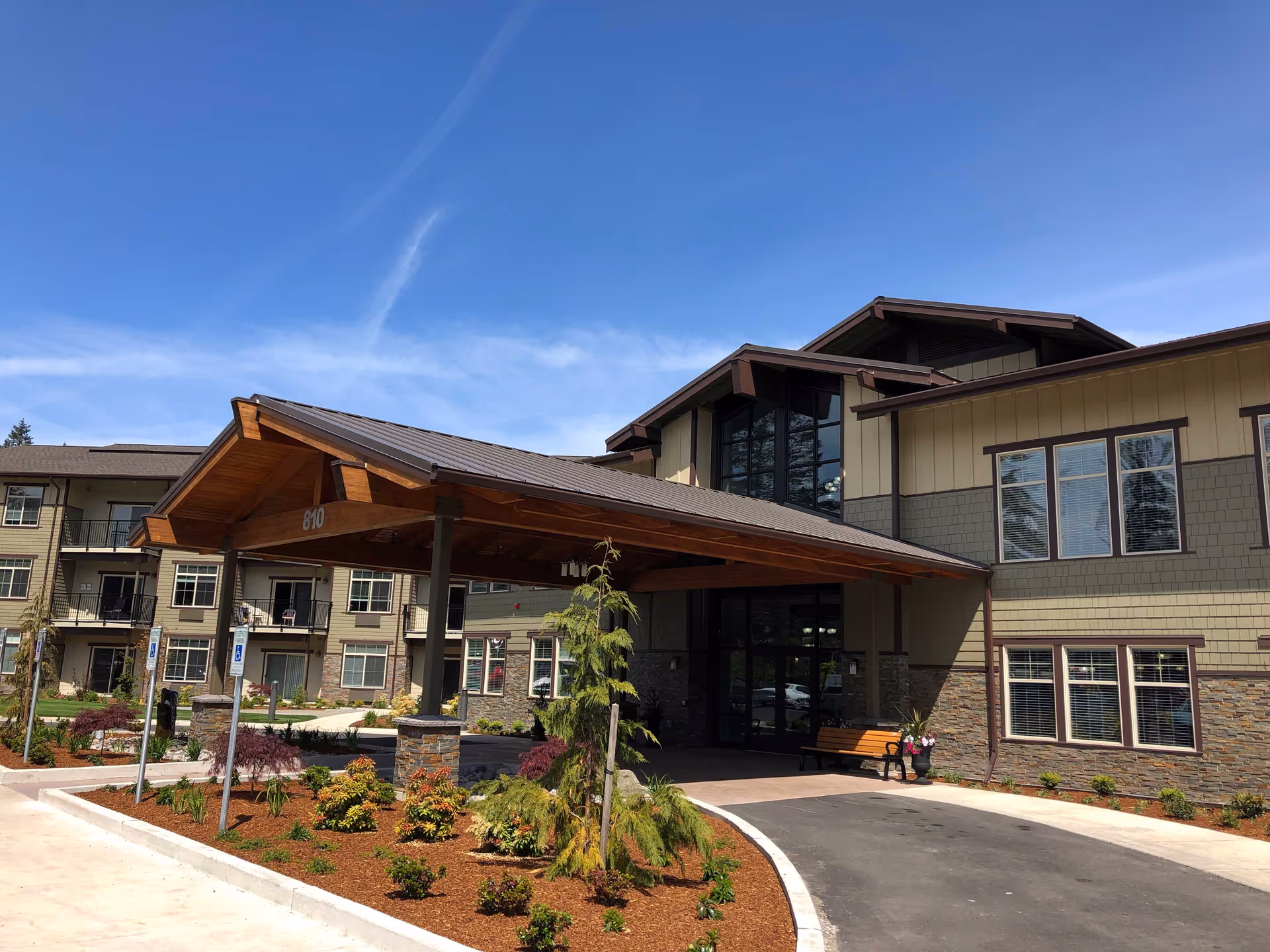 Exterior view of Fieldstone Cooper Point senior living facility showing the main entrance with a large wooden canopy, landscaped garden beds, and a clear blue sky.