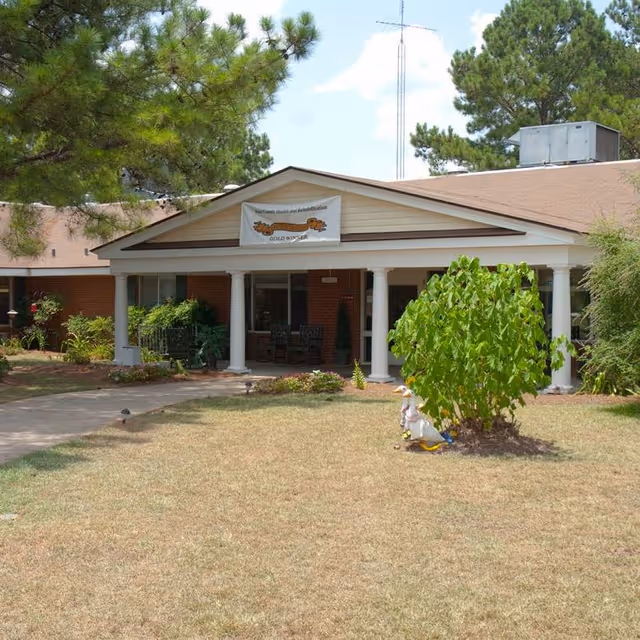 Front exterior view of a single-story brick building with white columns supporting a covered porch. There is a banner hanging above the porch that reads 'Four County Health and Rehabilitation GOLD WINNER'. The lawn in front has a small bush and a decorative duck statue. Trees and shrubs surround the building under a partly cloudy sky.