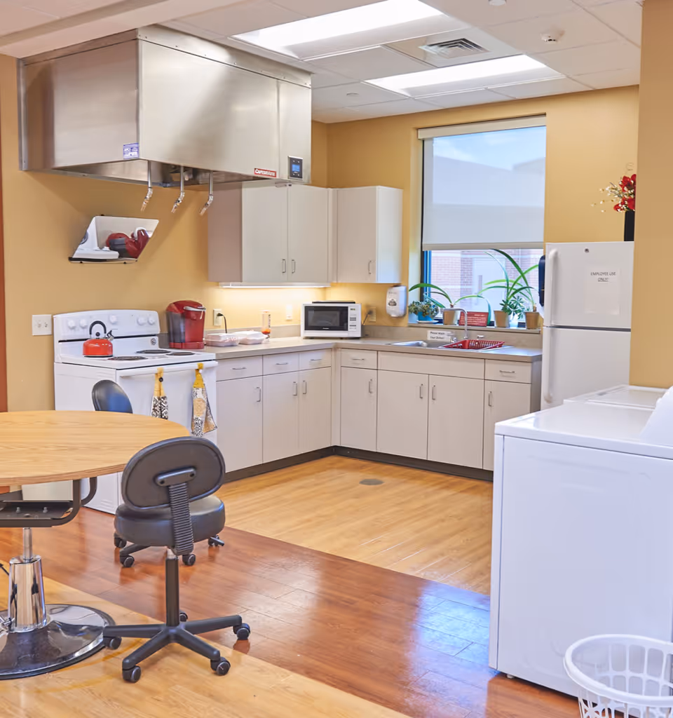 A clean and well-lit kitchen area with beige walls and wooden flooring. The kitchen features white cabinets, a white stove with a red kettle, a microwave, a red coffee maker, and a white refrigerator with a sign that reads 'EMPLOYEE USE ONLY'. There is a round wooden table with a black office chair on wheels. A window with a roller shade lets in natural light, and several potted plants are placed on the windowsill. A white washing machine and laundry basket are visible in the foreground.