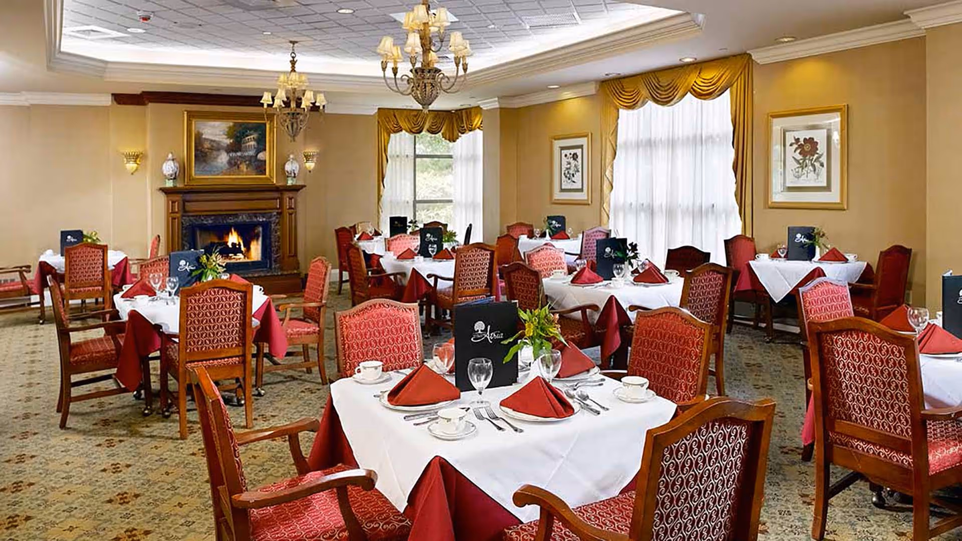 Elegant dining room with multiple tables covered in white and burgundy tablecloths, set with glassware, plates, and folded napkins. The room features red upholstered chairs, large windows with gold curtains, chandeliers, framed artwork on the walls, and a lit fireplace with a painting above it.