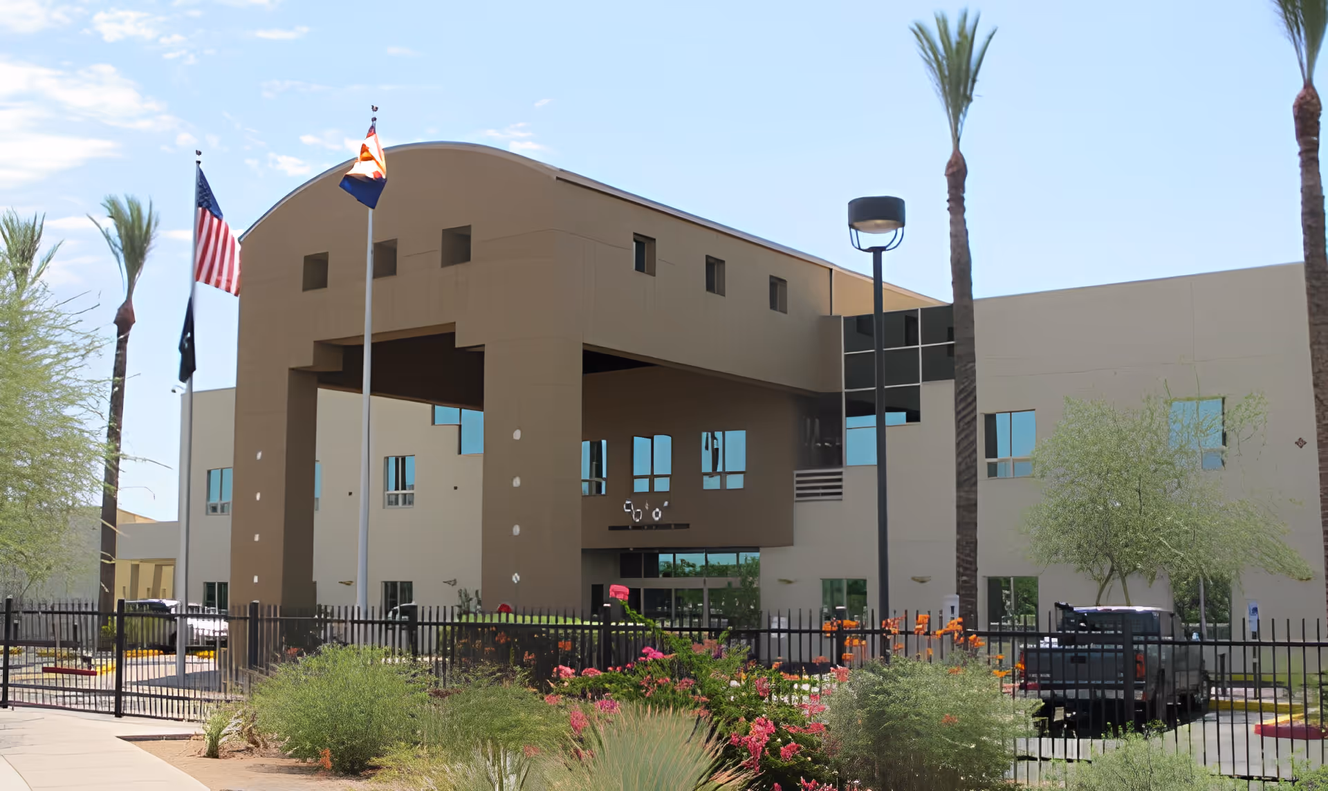 Exterior view of the Arizona State Veteran Home in Phoenix, showing a modern two-story building with a large covered entrance, three flagpoles with flags, palm trees, and landscaped greenery in the foreground.