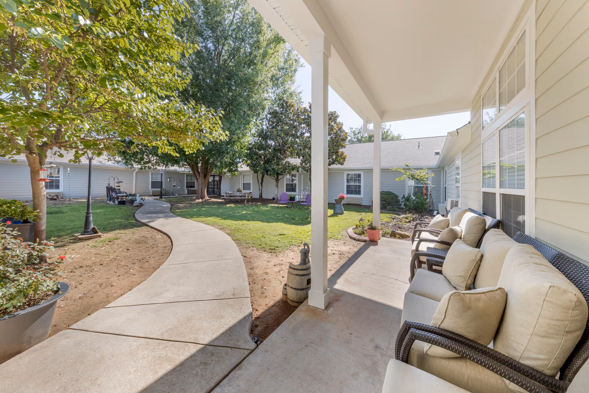 Covered porch with cushioned chairs overlooking a landscaped courtyard with a winding concrete path and trees.