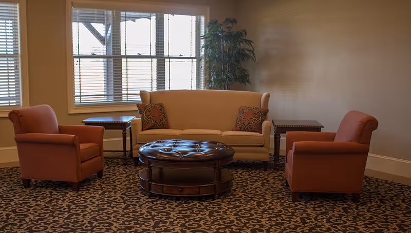 A cozy sitting area with a beige sofa adorned with two patterned cushions, two rust-colored armchairs, a round dark brown tufted ottoman, two wooden side tables, and a tall green plant near a window with blinds.