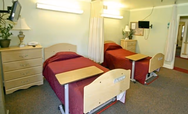 A shared patient bedroom with two hospital-style beds covered in burgundy bedspreads, bedside dressers and lamps, a wall-mounted TV, and a privacy curtain between the beds.