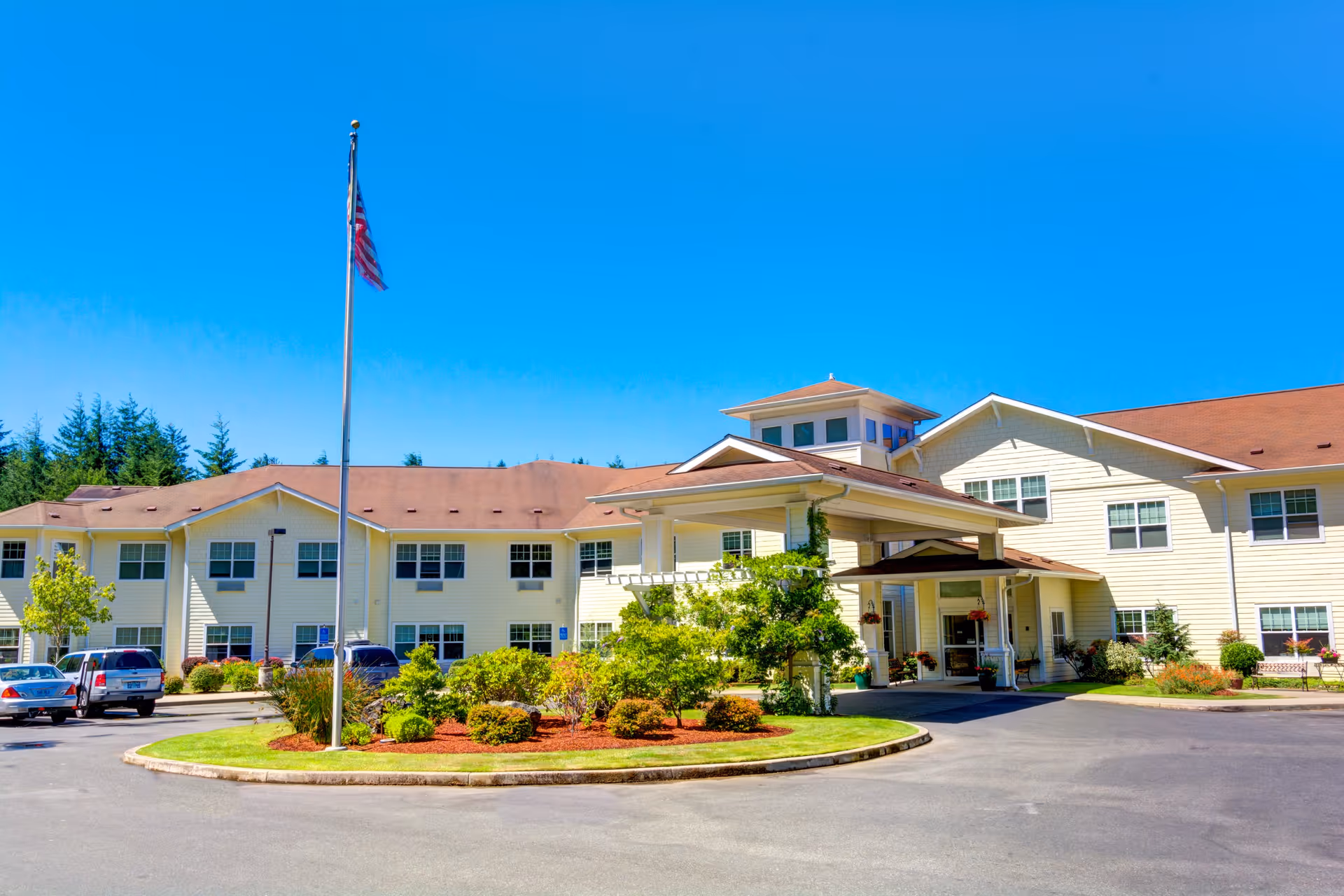 Exterior view of a two-story senior living facility building with beige siding and a brown roof under a clear blue sky. There is a circular driveway with landscaped greenery and an American flag on a flagpole in front of the entrance. Several parked cars are visible on the left side.