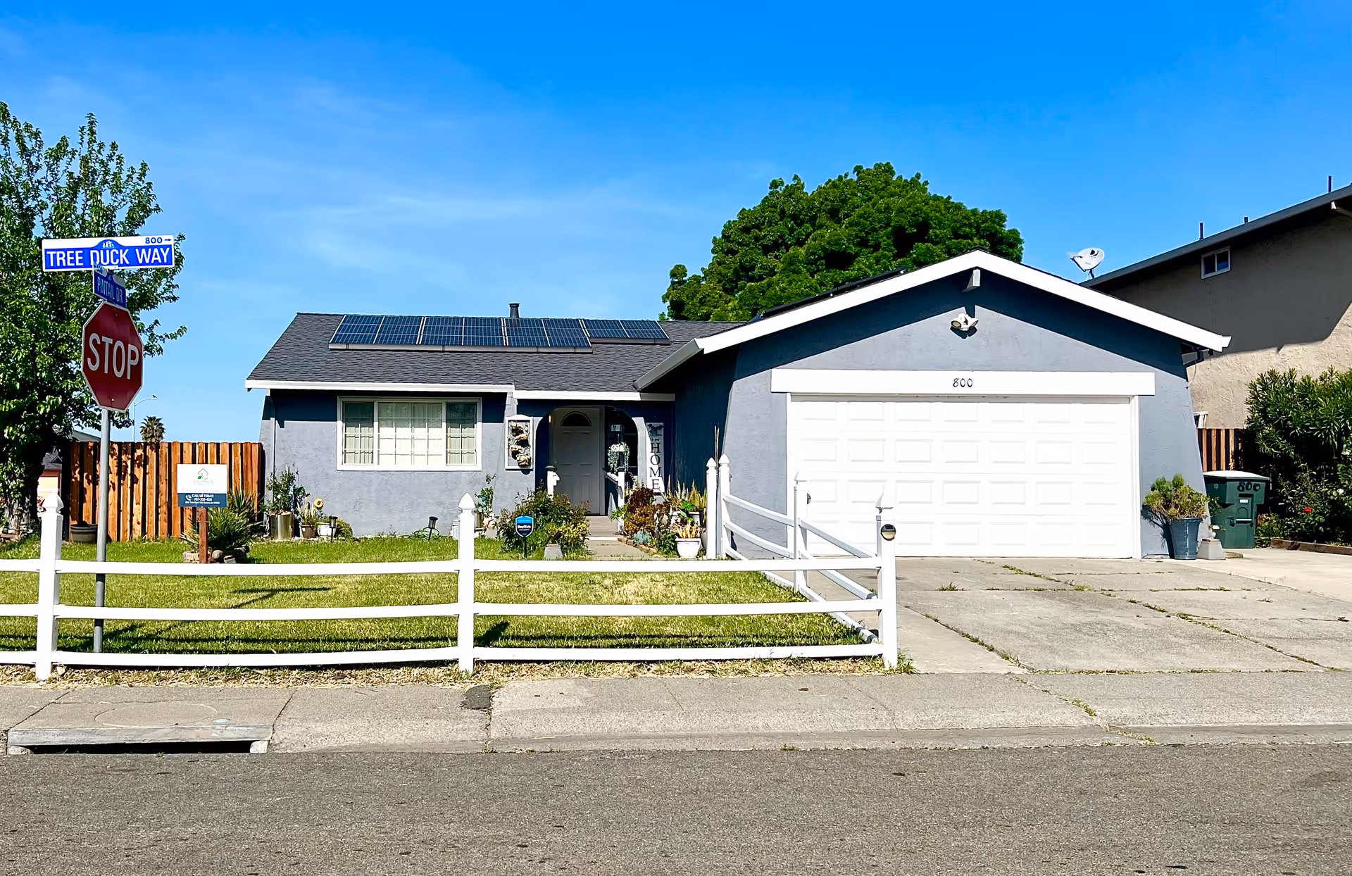 Single-story residential house with a gray exterior, white garage door, and solar panels on the roof. The front yard has a green lawn, white fence, and various plants. A street sign reads 'Tree Duck Way' next to a stop sign. The sky is clear and blue.