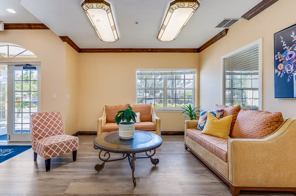 A cozy senior living facility lounge area with two beige sofas adorned with patterned and solid-colored cushions, a patterned armchair, and a round coffee table with a potted plant. The room has large windows letting in natural light, wooden crown molding, and two decorative ceiling lights.