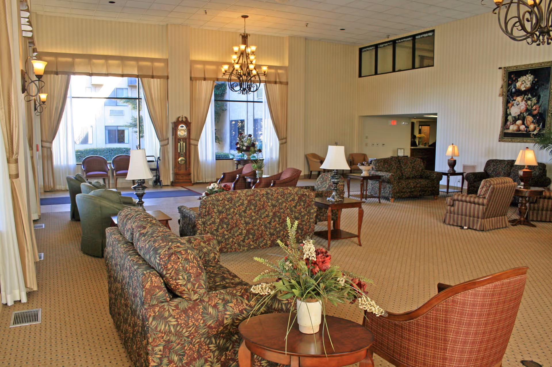 Spacious furnished lounge in a senior living facility with patterned sofas, armchairs, lamps, chandeliers, and large windows.