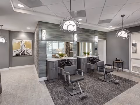 Interior view of a modern hair salon area within a senior living facility, featuring two black salon chairs in front of two large mirrors mounted on a gray textured wall. The space is well-lit with geometric pendant lights hanging from the ceiling. There is a small table with a phone and a decorative plant, and a hallway leading to other rooms with artwork on the walls.