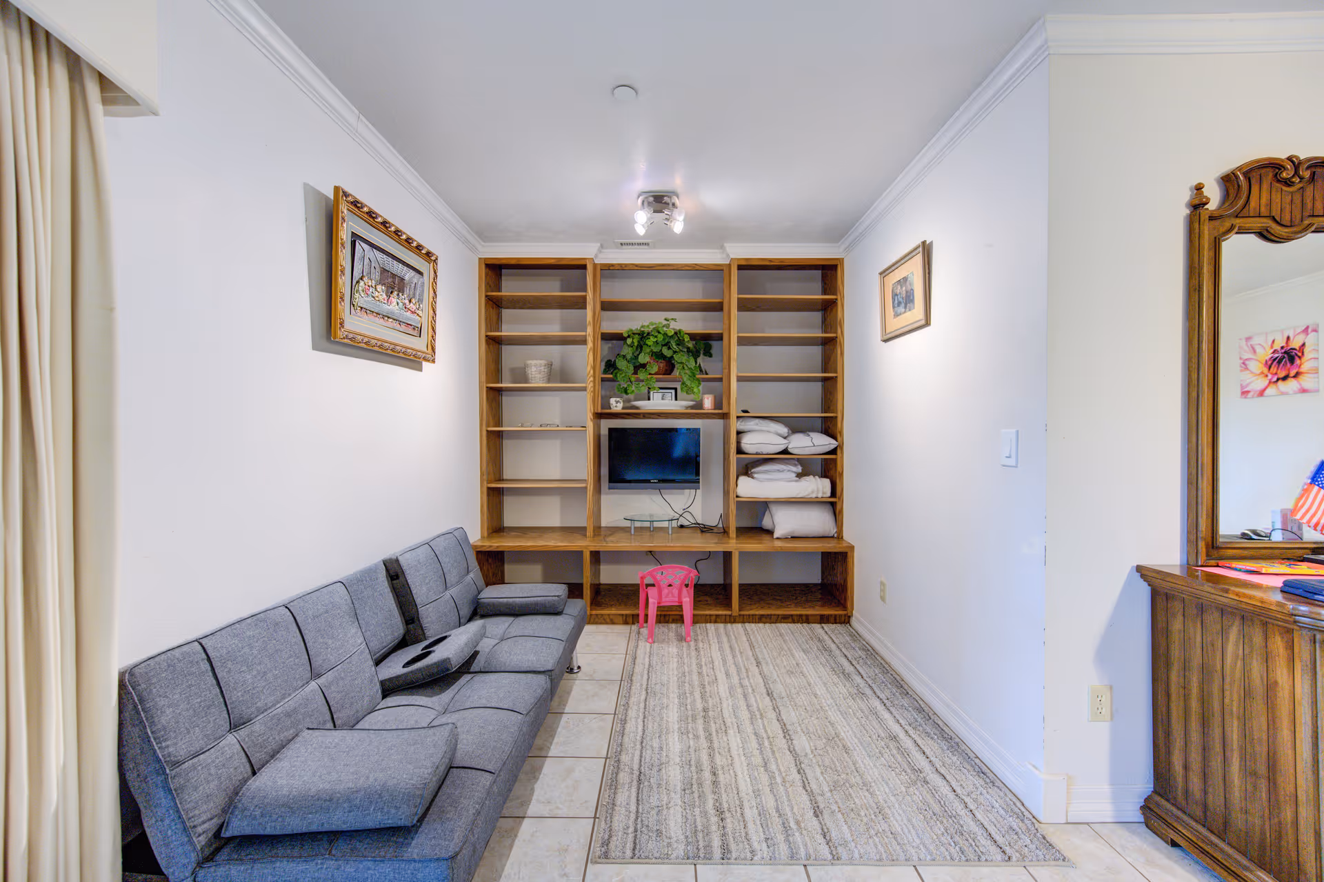 Small living room with a gray sofa, built-in wooden shelving holding a small TV and folded linens, a rug, and a wooden dresser with mirror.