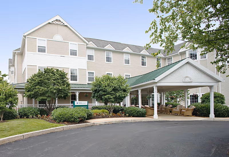 Exterior view of Highgate at Paoli Pointe, a multi-story senior living facility with beige siding and white trim. The building features multiple windows and a covered entrance with a green roof. There are trees, shrubs, and landscaped greenery around the entrance area, and a paved driveway in front.