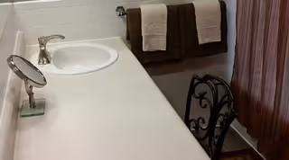 A bathroom countertop with a white sink, a silver faucet, a small round mirror on a stand, and two brown towels hanging on a towel rack against a white tiled wall. There is a decorative black metal chair with a cushion next to the counter and a brown shower curtain in the background.