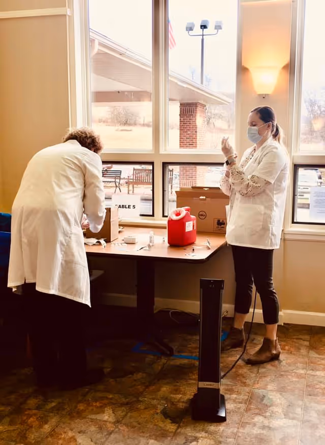 Two healthcare workers wearing white coats and face masks preparing medical supplies on a table near large windows with an outdoor view. One person is standing and handling a syringe, while the other is bent over the table organizing items.